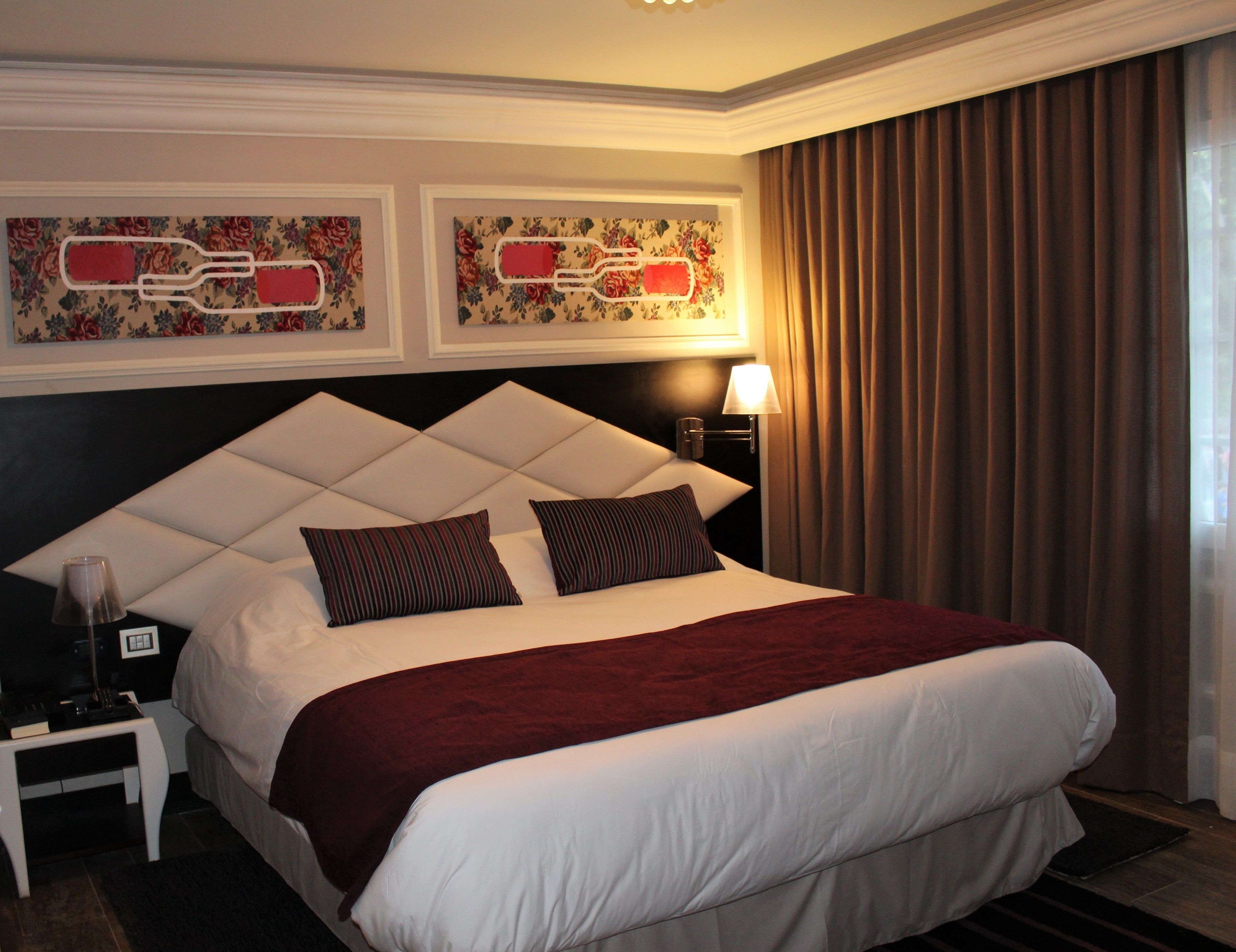 Una habitación de hotel con una cama grande, una mesita de noche, una lámpara y dos cuadros con motivos florales en la pared.