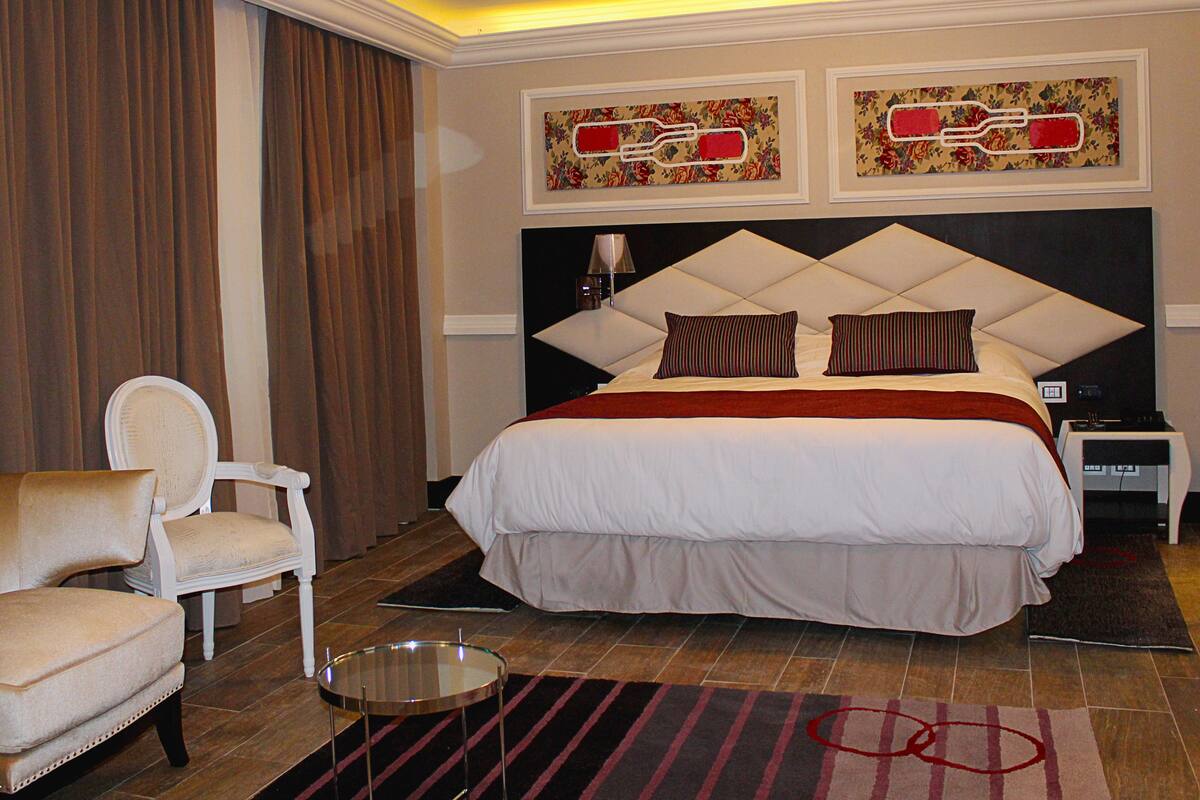 A hotel room with a large bed, a chair, a bedside table, and artwork on the wall.