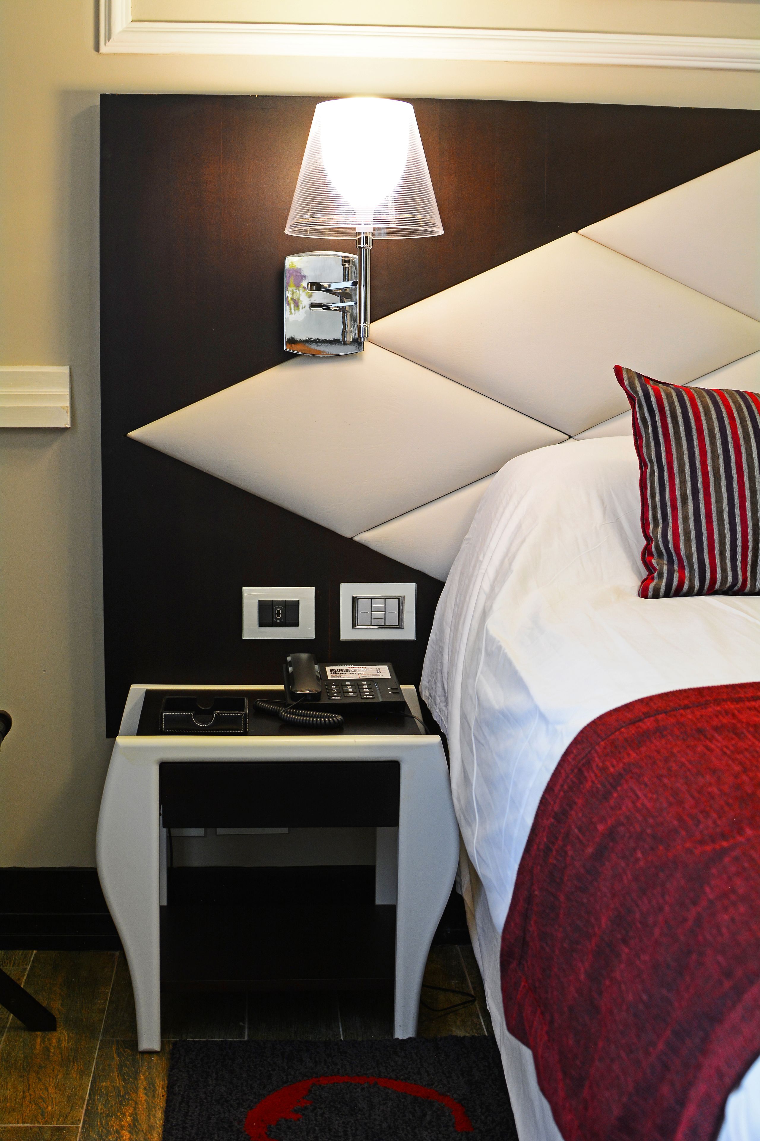 Habitación de hotel con cama, mesita de noche con lámpara, teléfono y panel de control en la pared.