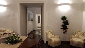 En matplats med ett träbord, stolar och en vas med blommor. Det finns två fåtöljer och en krukväxt. En hall leder till ett annat rum.