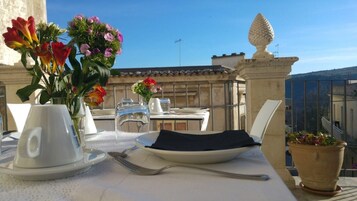 Mesa para dos con un jarrón de flores, una taza y un plato en un balcón con vista a la ciudad.