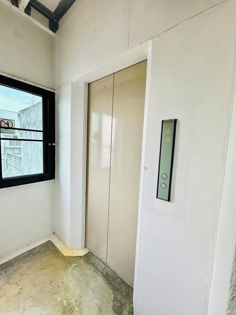Ein schmaler Flur mit einer zur Kammer führenden Tür, einem Fenster mit Blick auf ein Gebäude und einem an der Wand befestigten Bedienfeld für den Aufzug.
