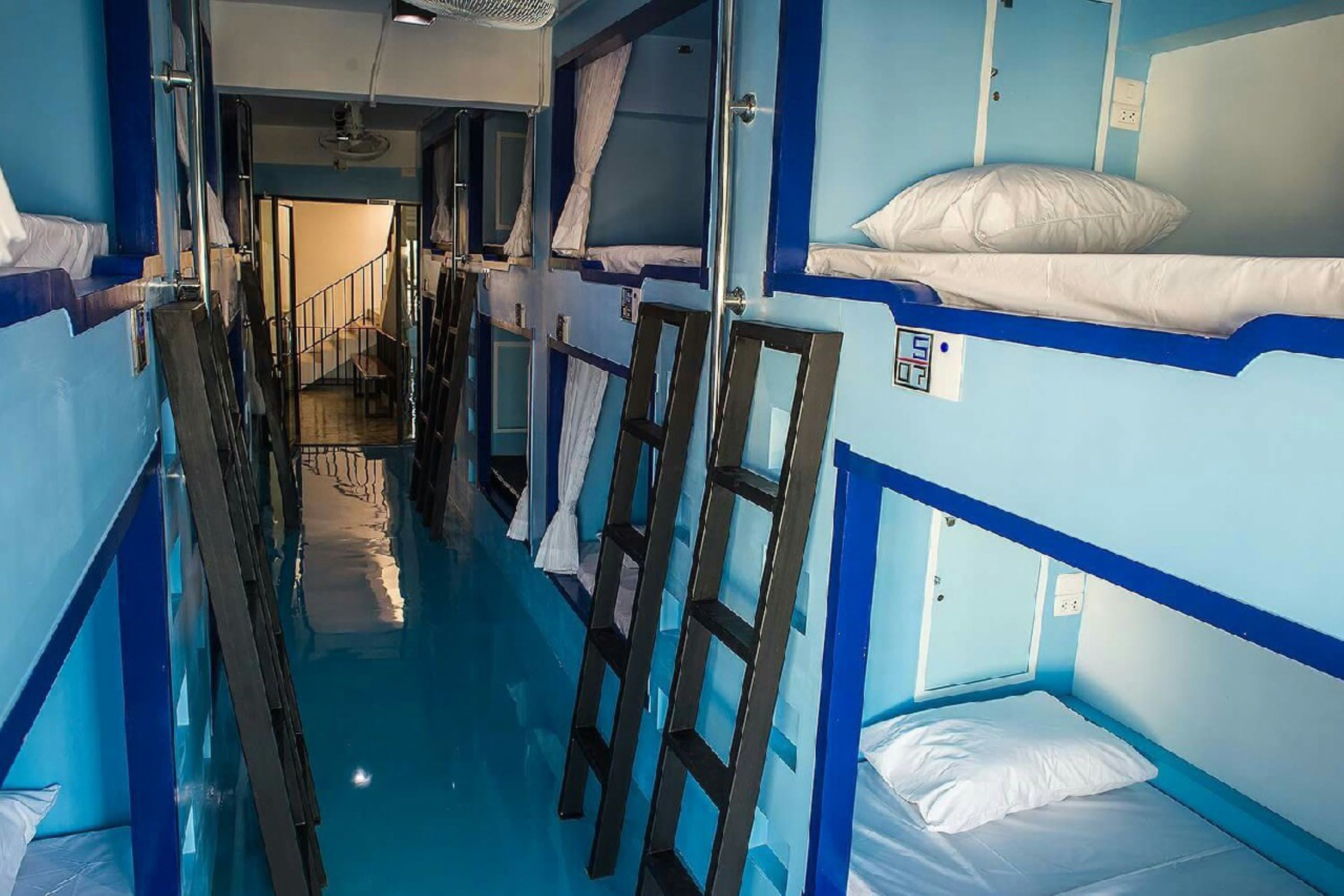 A narrow corridor with bunk beds on either side, a visible staircase, and a ceiling fan.