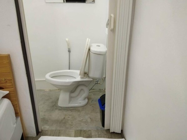 A small, narrow bathroom with a white toilet, a visible pipe, and a blue trash bin. - The Port Hostel By Suwatchai (Surat Thani)