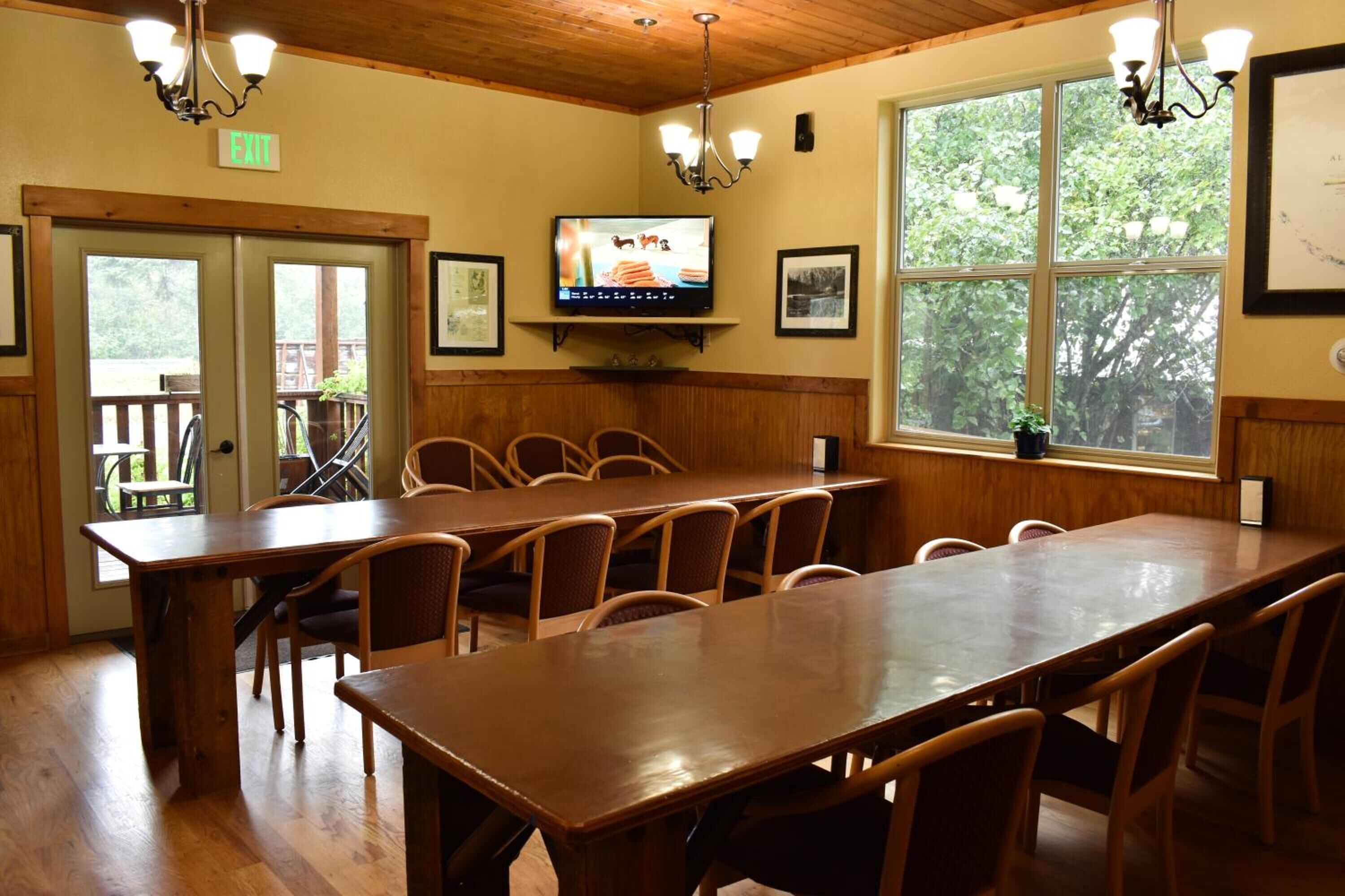A conference room with a long table and chairs, a television on the wall, and large windows with a view of trees.