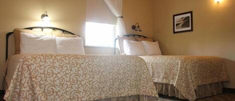 A hotel room with two beds, each with a patterned bedspread, a window with a white curtain, and a wall-mounted picture.
