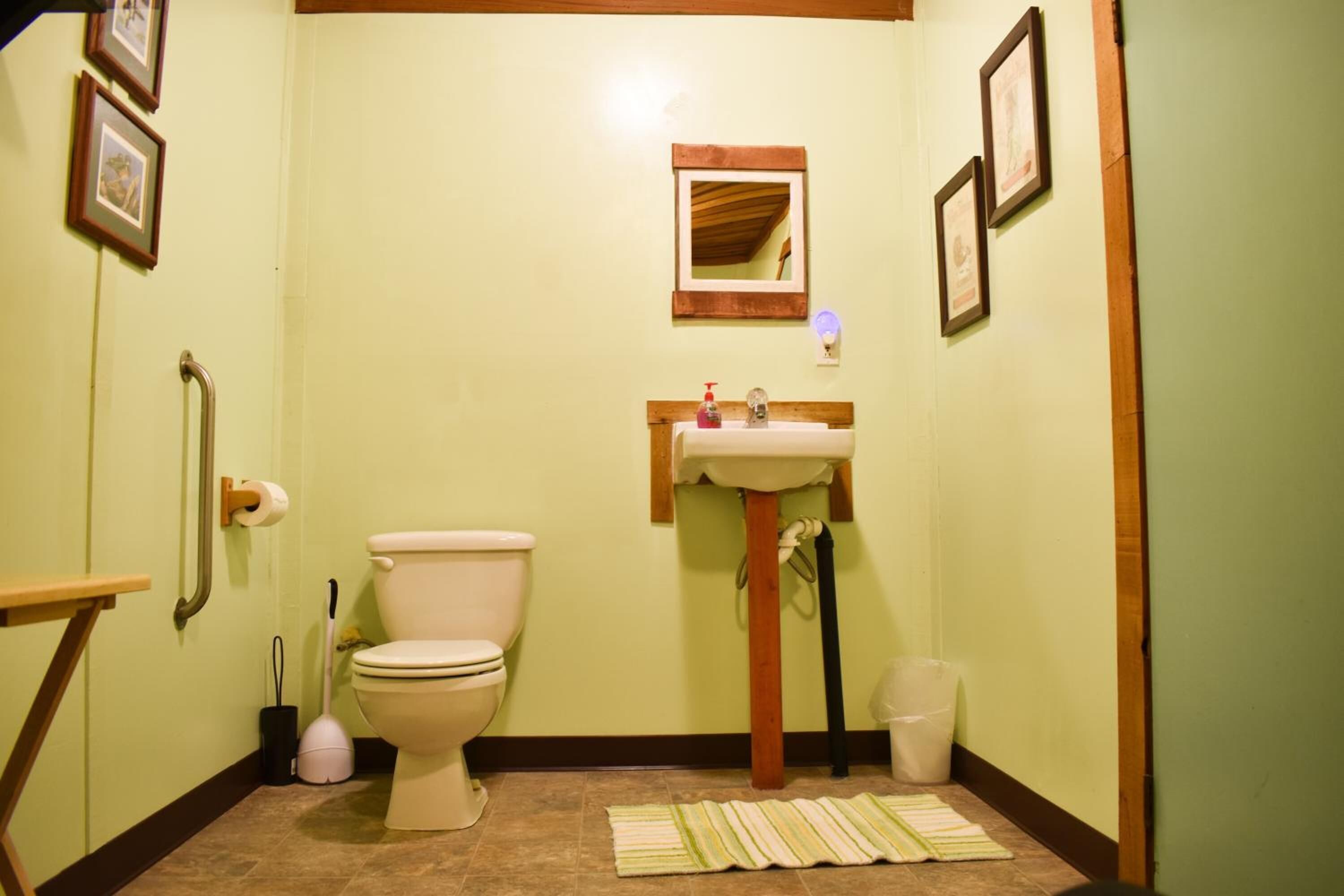 A small bathroom with a toilet, sink, and mirror. There is a handrail on the left and a trash can on the right.