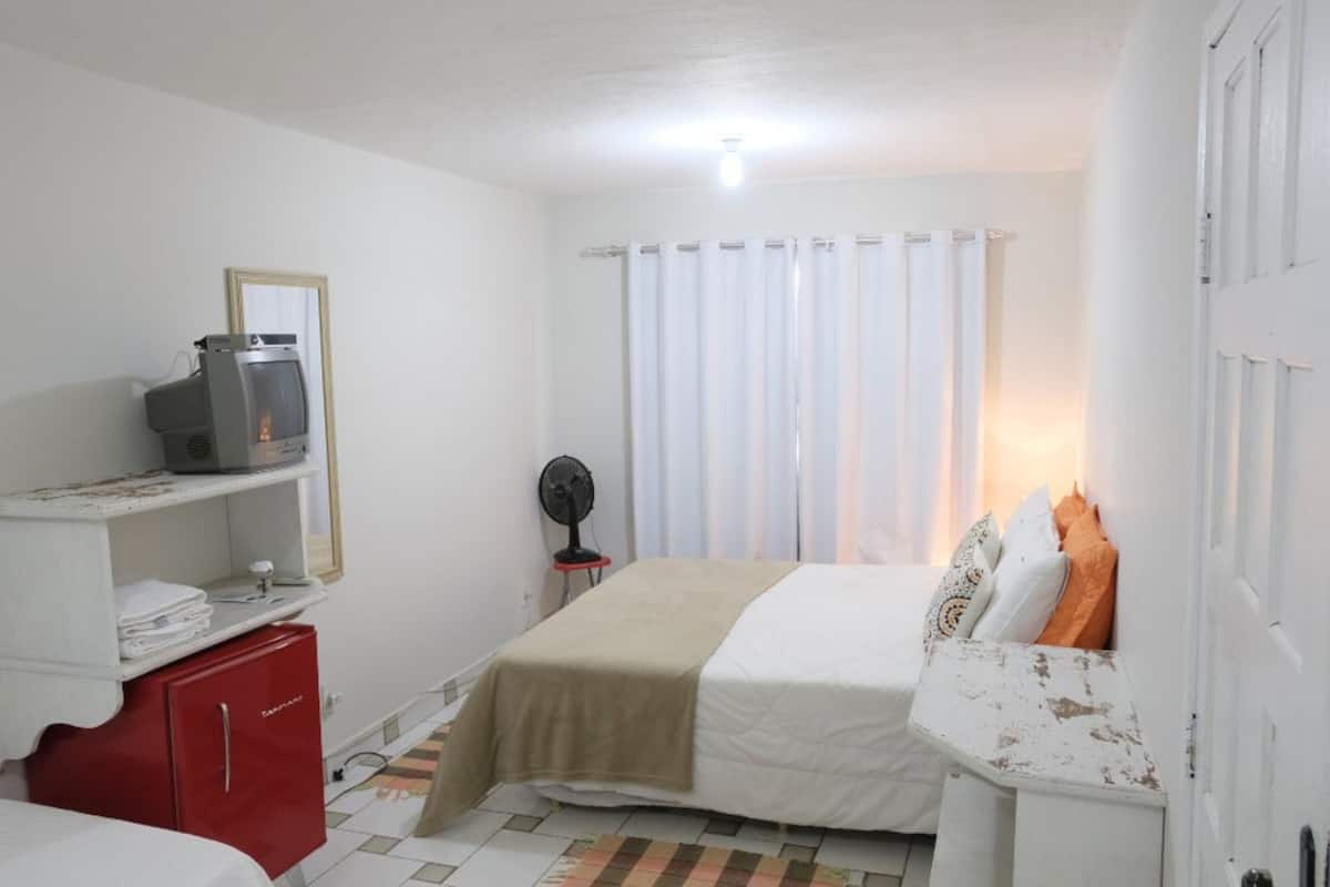 A small, clean room with a bed, a red cabinet, a television, and a fan.