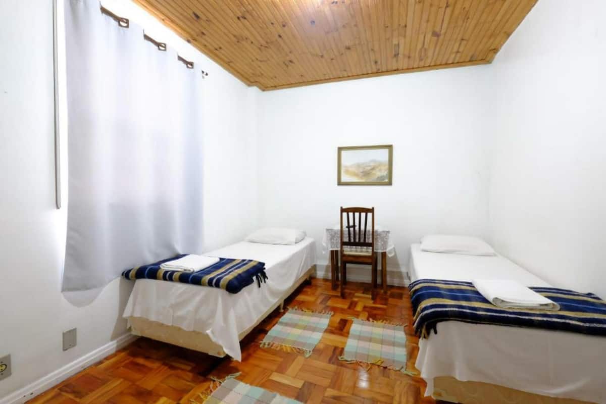 A small, clean room with two single beds, a wooden ceiling, a table with a chair, and a painting on the wall.