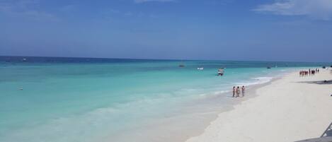 A beach with clear turquoise water, a sandy shore, and a few boats in the distance.