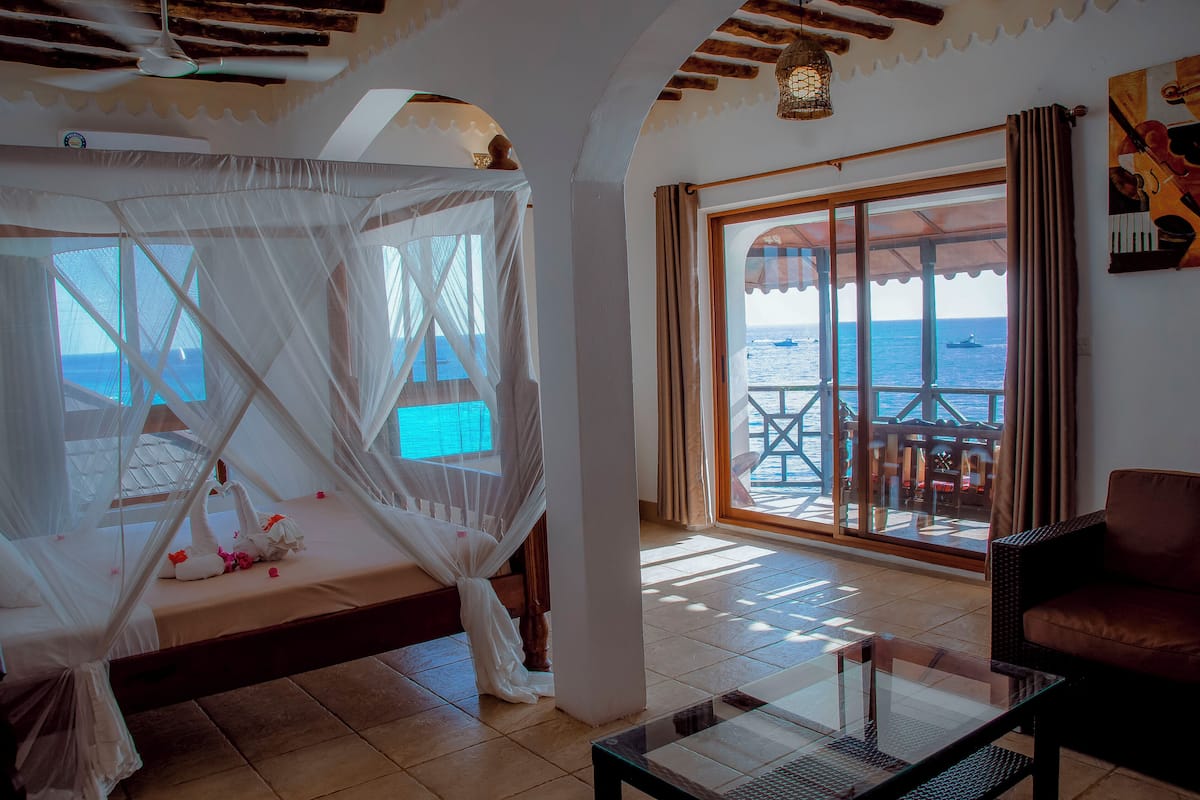 Une chambre avec un lit sous un dais blanc, une table en verre, un canapé marron et une vue sur la mer grâce à de grandes fenêtres.