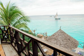 Eine Holzterrasse mit einem Reetdach, mit Blick auf türkisfarbenes Wasser und ein Segelboot.