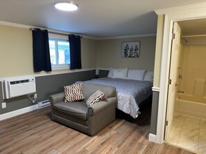 A bedroom with a bed, a sofa, a window with curtains, a radiator, and a bathroom door. - Sunset Motel at Armand's Gardens  (St Ignace)