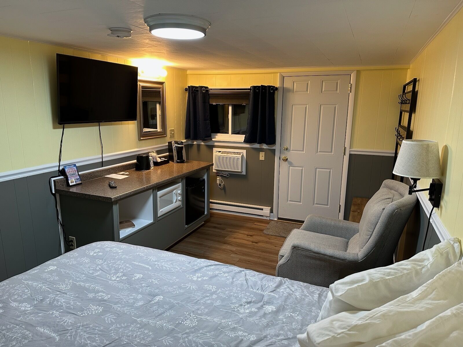 Ein Hotelzimmer mit Bett, Schreibtisch, Flachbildfernseher, Stuhl, Lampe und Tür.