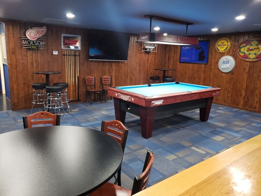 A pool table in a room with wooden walls, a bar area, and a television mounted on the wall.