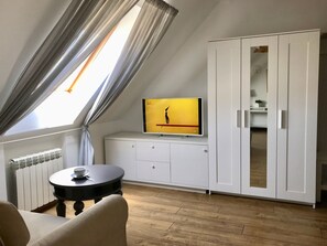 A room with a sloped ceiling, a TV on a white cabinet, a round table with a cup, and a wooden floor. - Hotel Address (Saratov)