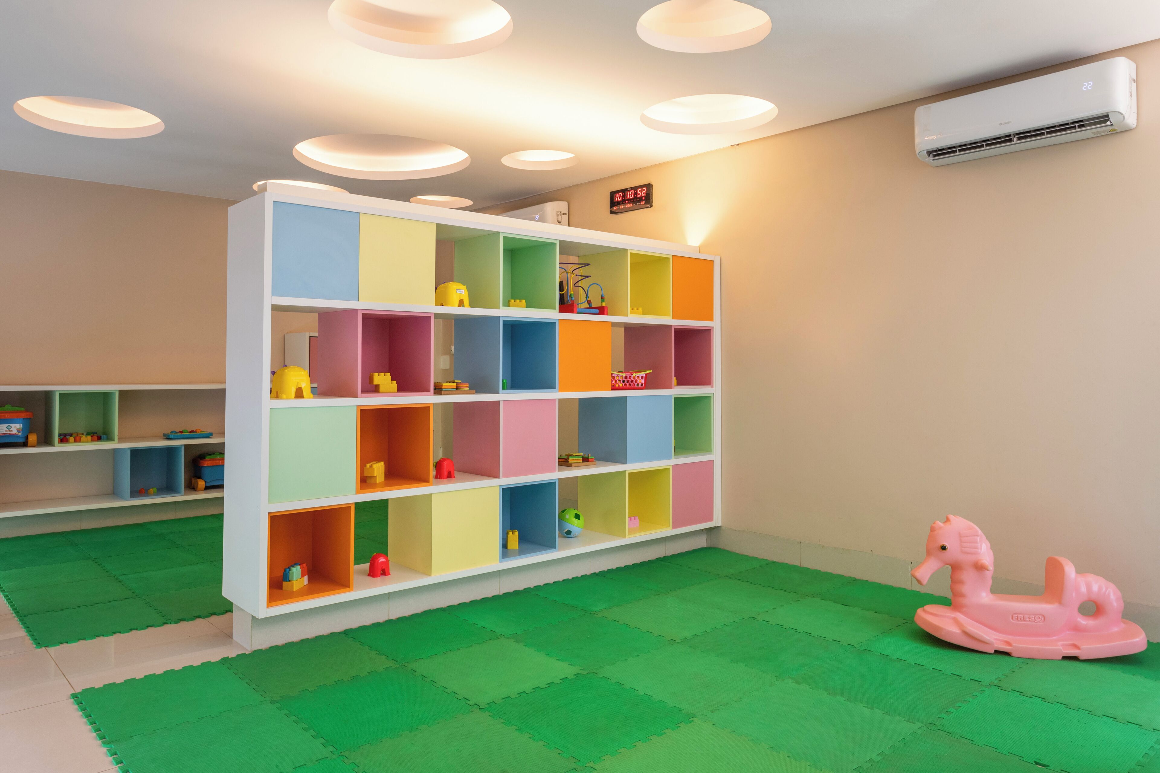 Quarto com uma estante colorida, um tapete de brincadeira verde e um cavalo de brinquedo rosa.