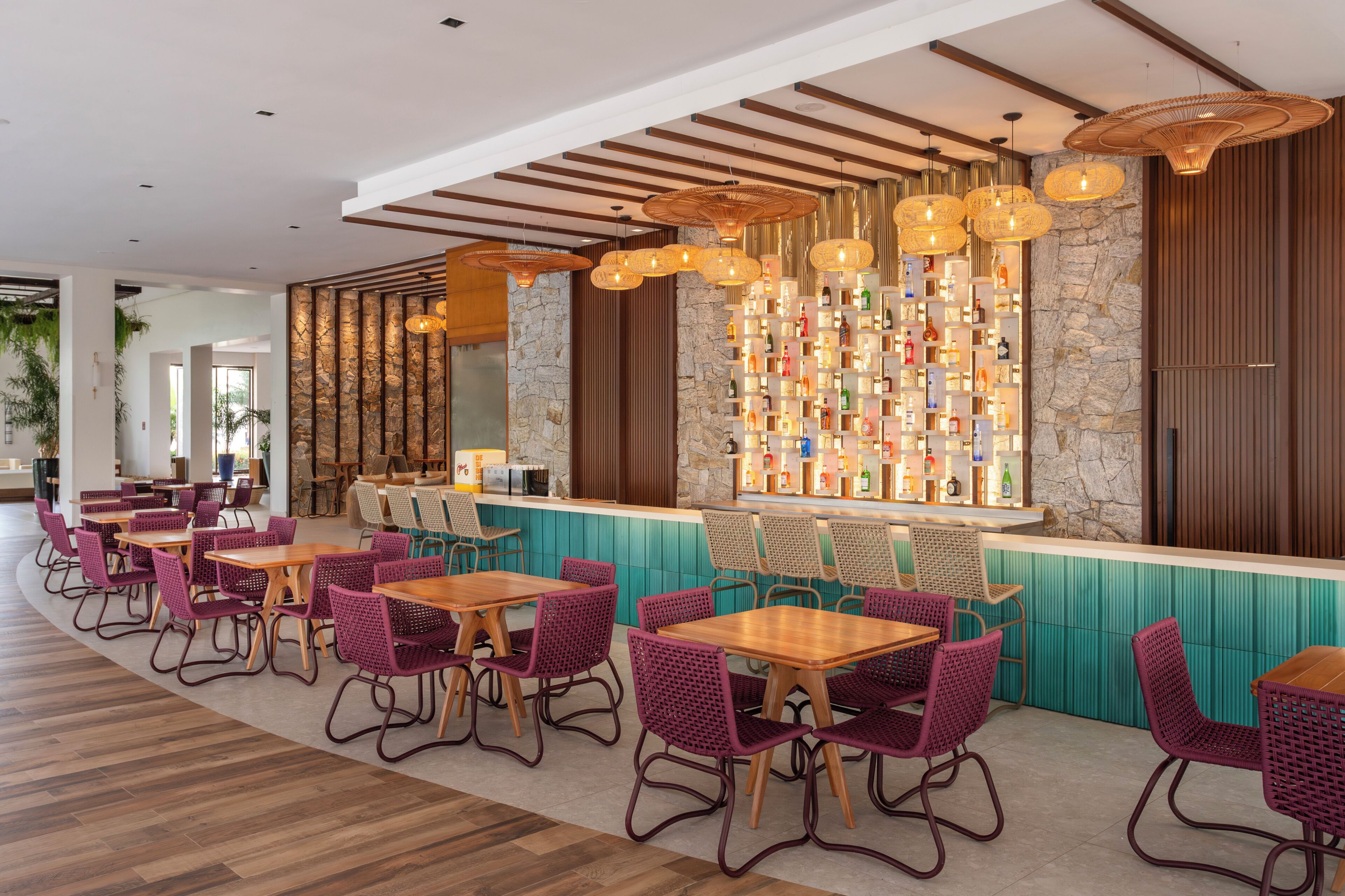 A restaurant with wooden tables and chairs, a stone wall with a bottle display, and hanging wicker lamps.