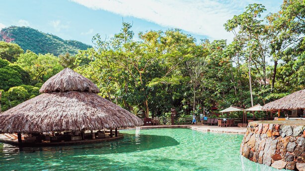 A clear pool with a thatched-roof structure, surrounded by lush greenery and a mountainous backdrop. - Rio Quente Resorts - Cristal Resort (Rio Quente)