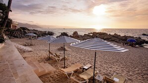 Een strand met ligbedden en parasols, een trap die naar het zand leidt, en een uitzicht op de oceaan en bergen in de verte.