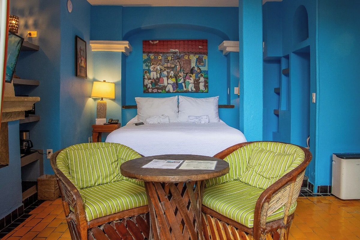 Ein Hotelzimmer mit einem großen Bett, zwei grün gestreiften Sesseln, einem kleinen runden Tisch, einem Bild an der Wand und einem Heizgerät am Boden.