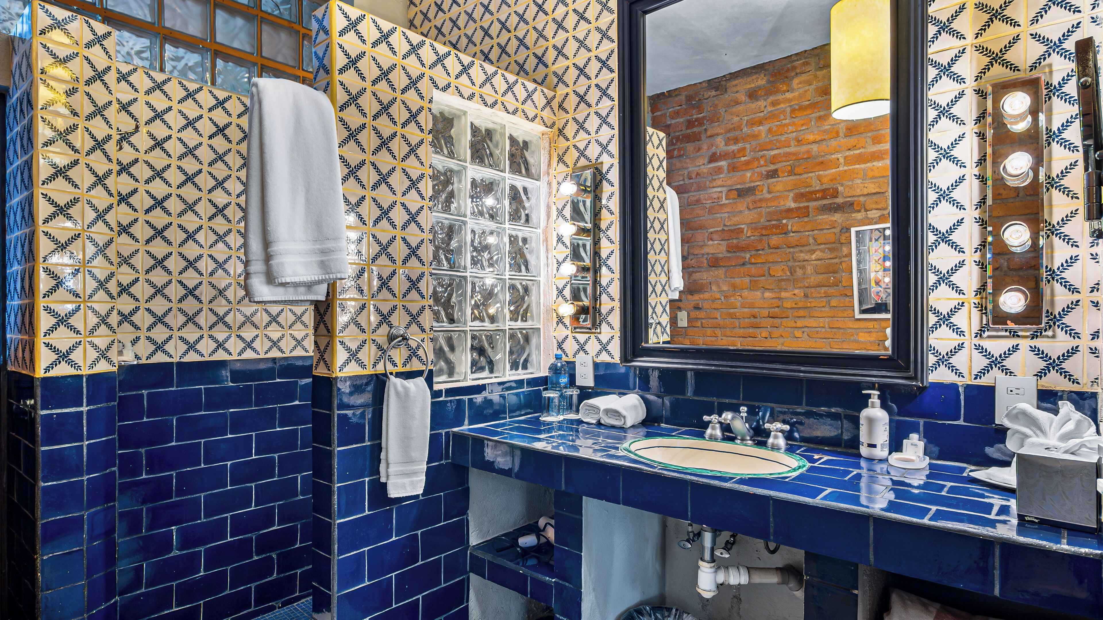 A bathroom with blue and yellow patterned tiles, a mirror with lights, a sink with toiletries, and a towel rack.