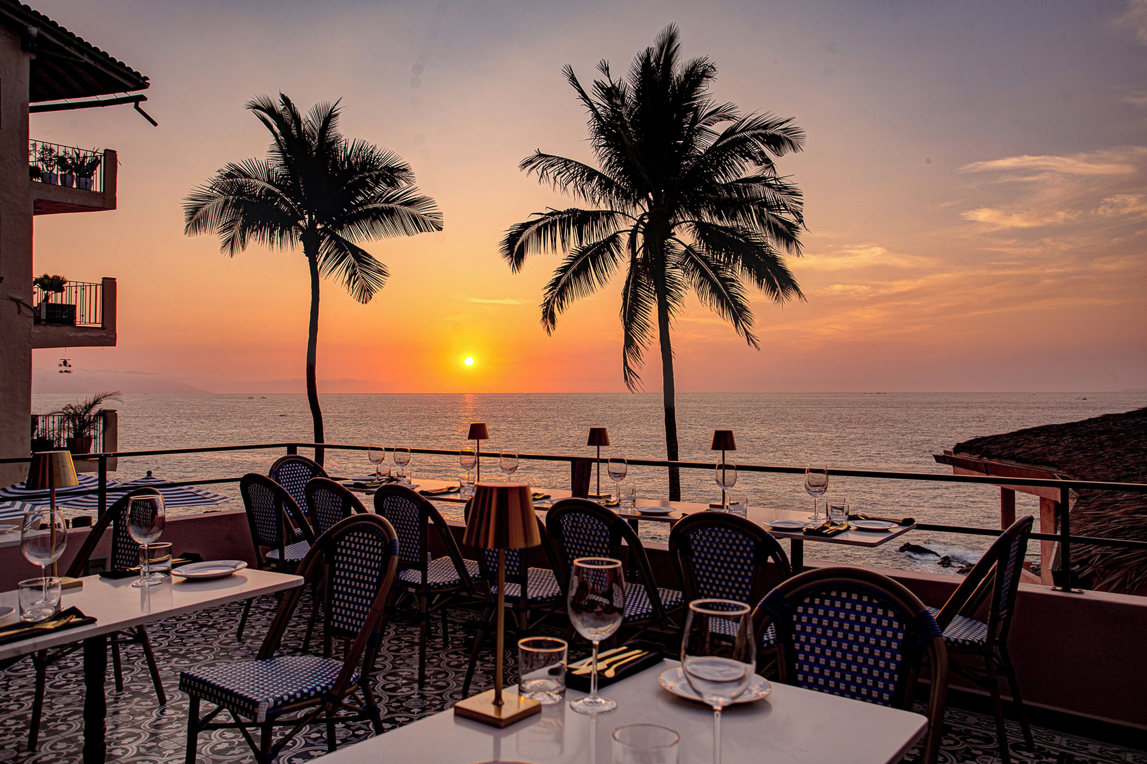 A restaurant with tables and chairs facing a sunset over the ocean, palm trees, and a balcony overlooking the sea.