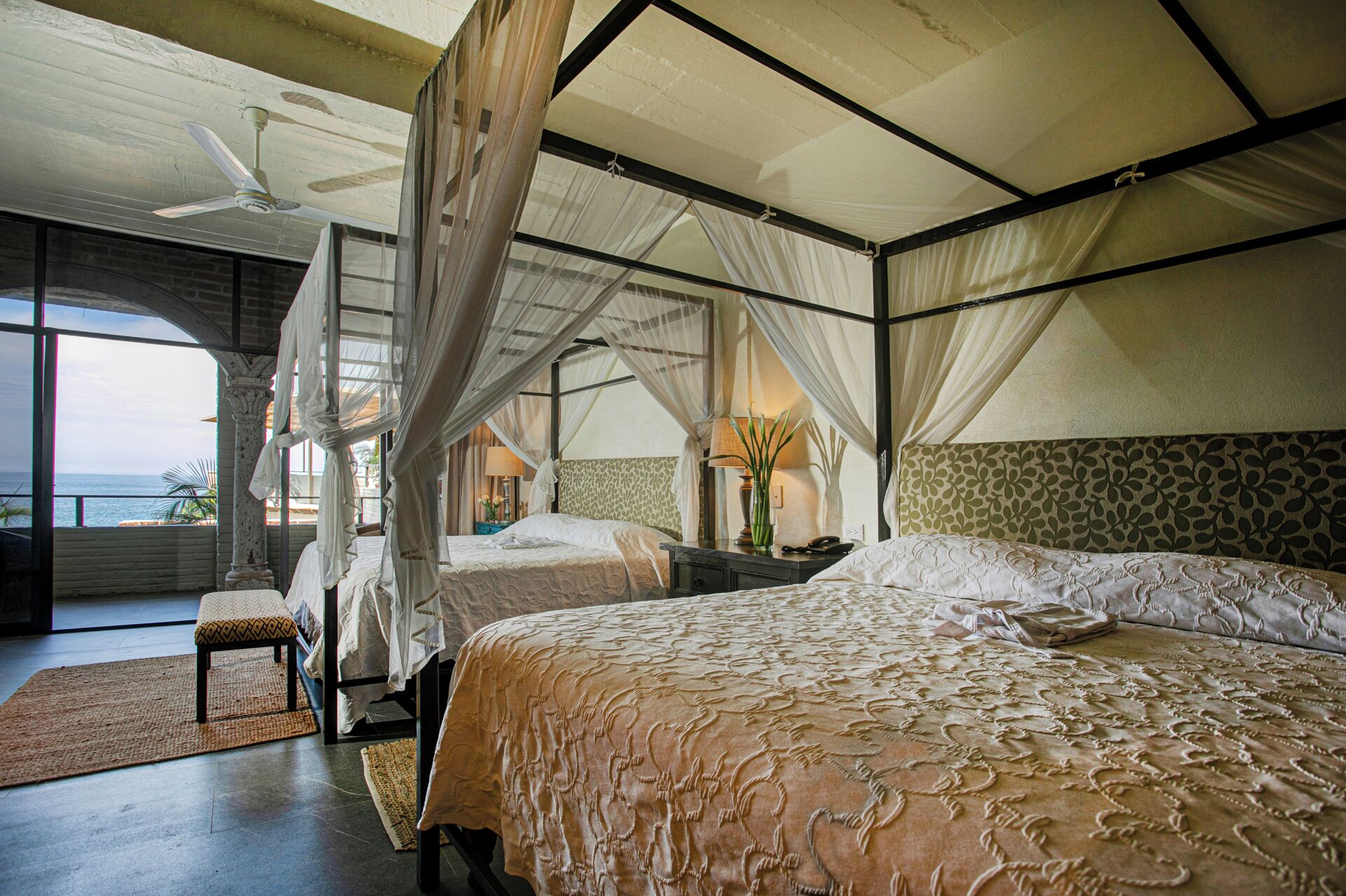 Ein Schlafzimmer mit einem großen Bett, einem kleineren Bett, einer Bank und einem Nachttisch. Ein Fenster bietet Blick auf den Ozean.