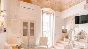 Ein Zimmer mit Steindecke, weißen Wänden, einem weißen Sessel, einem kleinen runden Tisch, einer weißen Kommode und einer Treppe.