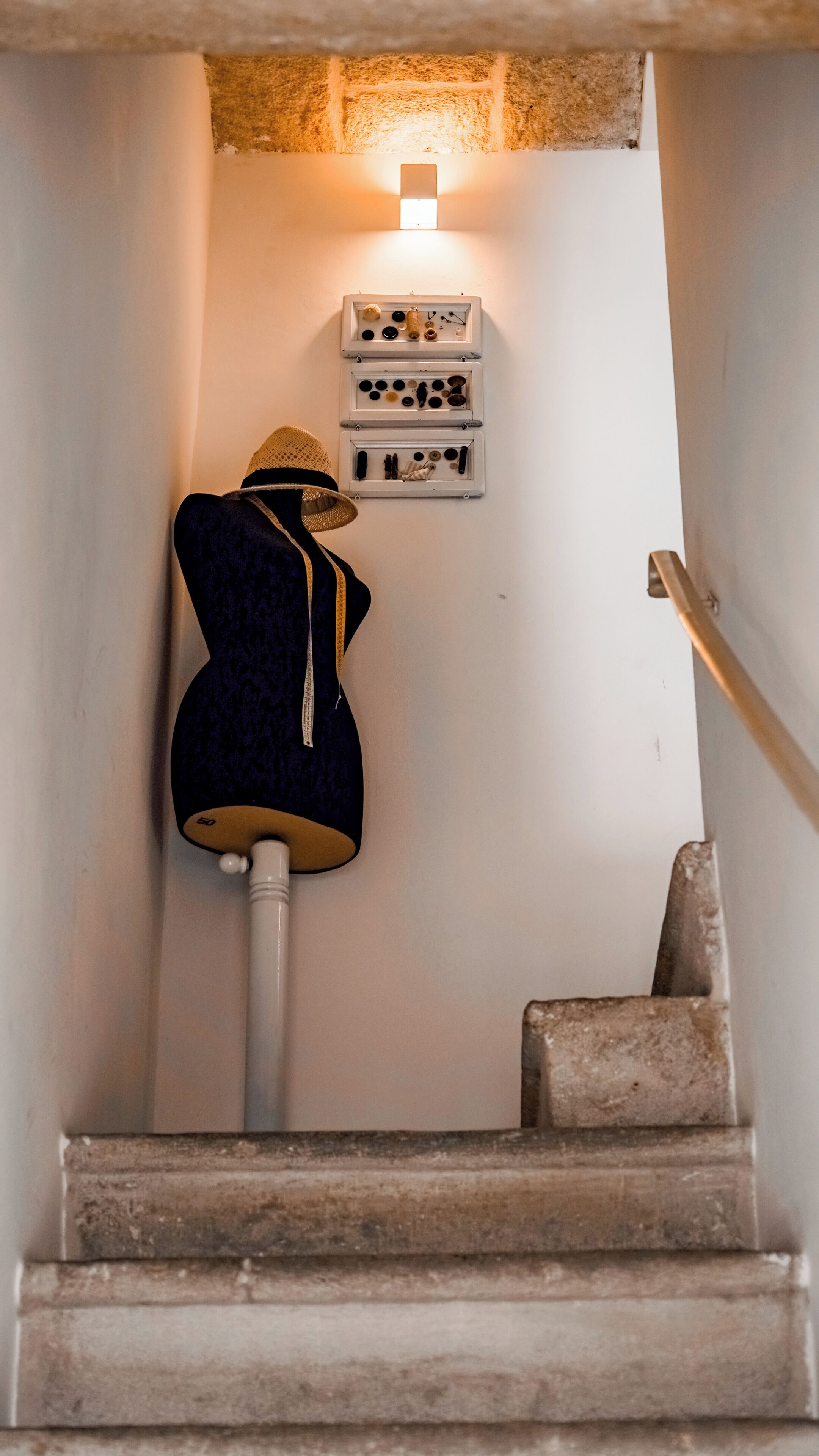 Un escalier avec un mannequin portant une tenue sombre, un luminaire fixé au mur et une étagère contenant divers objets.