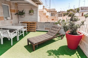 A rooftop terrace with a lounge chair, a table, and chairs, surrounded by potted plants and a red pot.