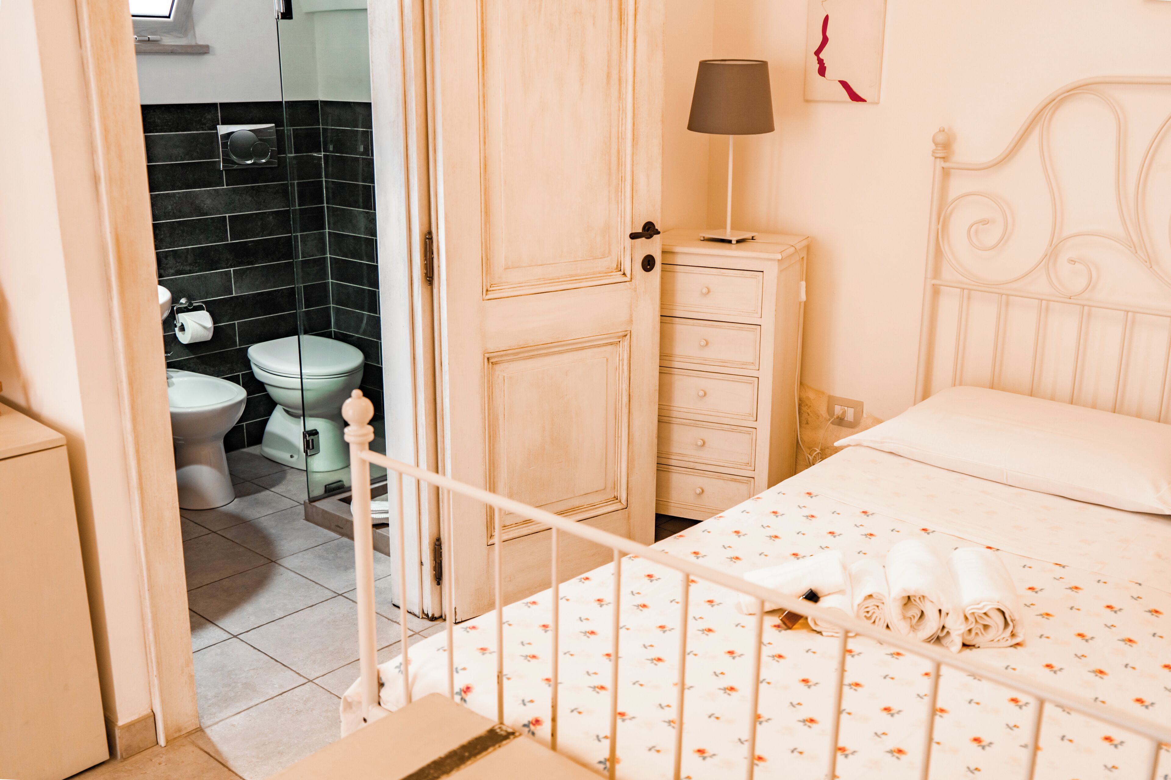 Quarto com cama, mesa de cabeceira, luminária e banheiro visível por uma porta aberta.