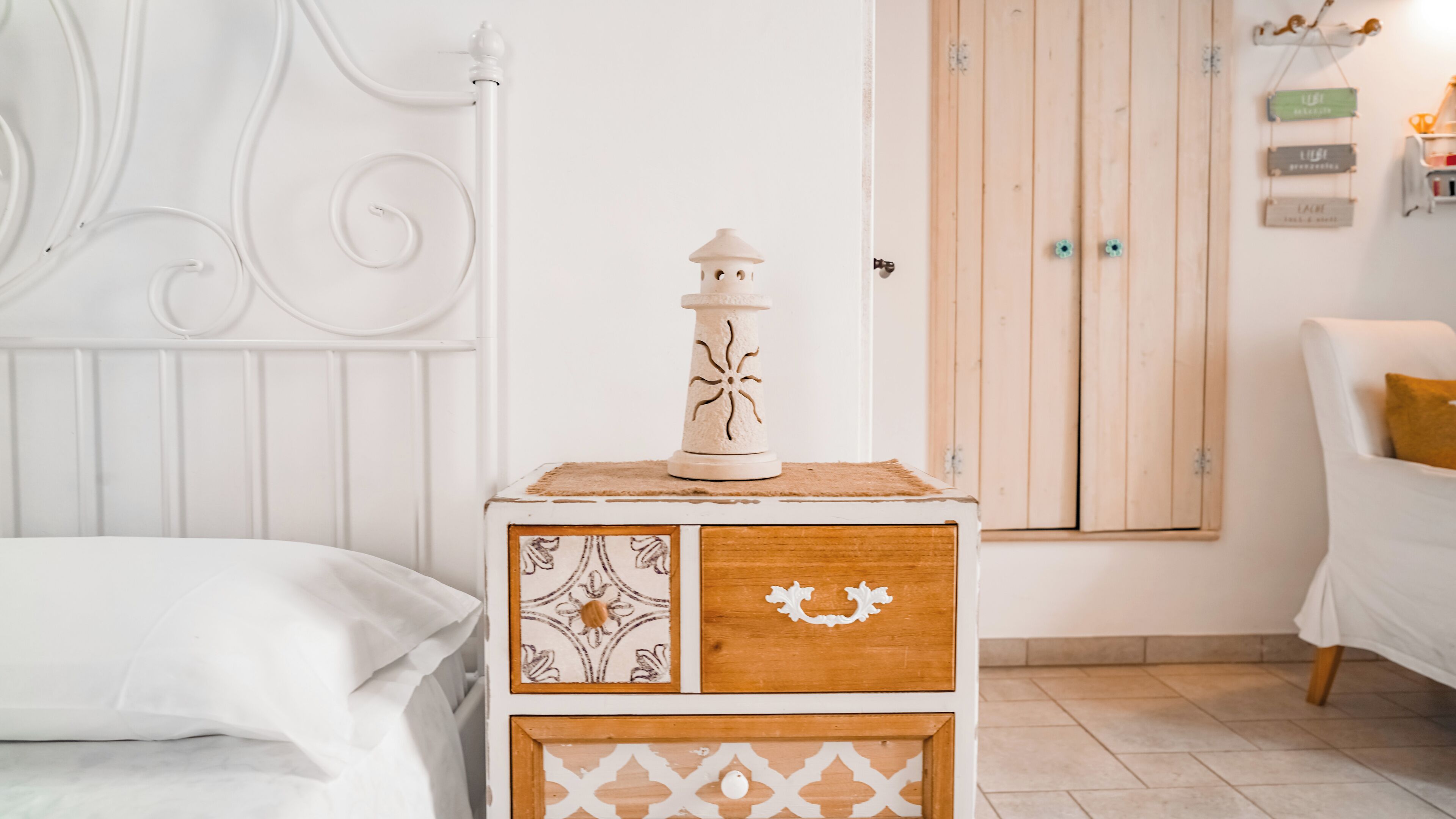 Una estructura de cama metálica blanca, una mesita de noche de madera con una lámpara decorativa y un armario de madera con puerta.