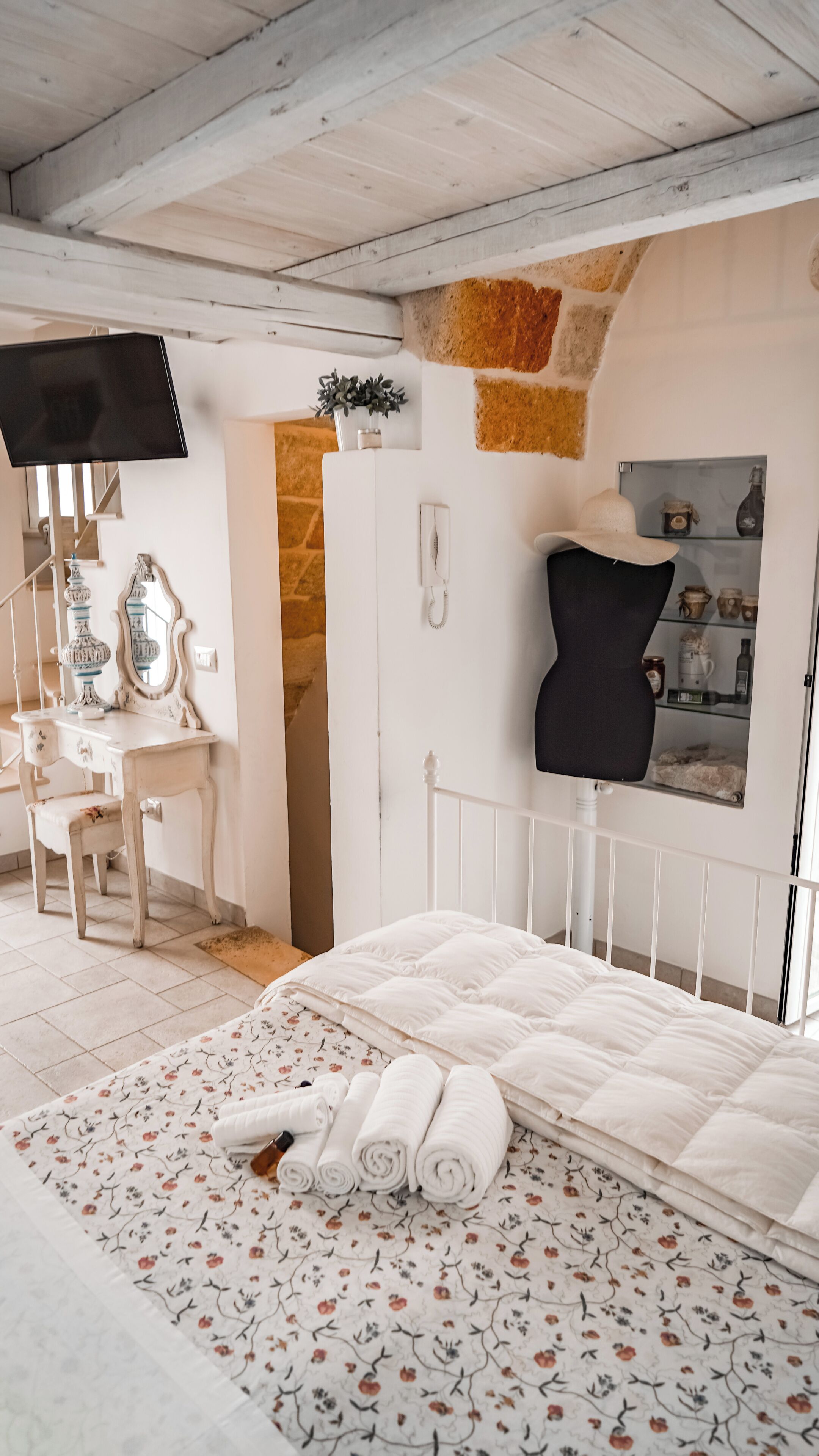Quarto com cama, escrivaninha, cadeira, televisão e prateleira na parede com diversos objetos.