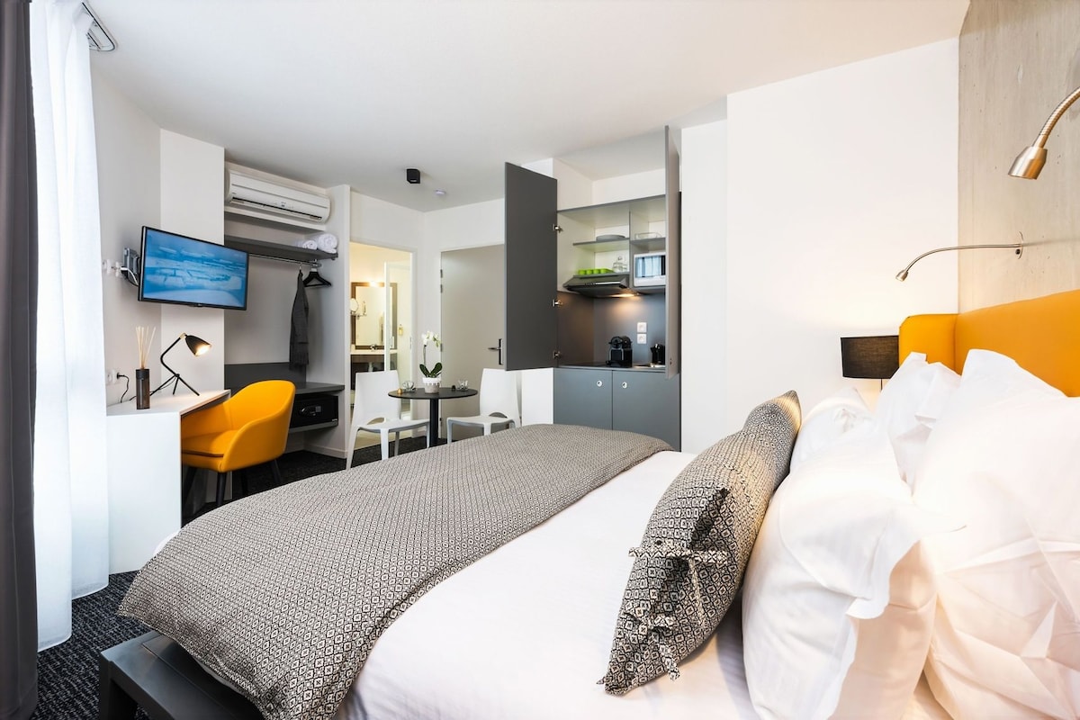 Habitación de hotel moderna con cama, escritorio, silla, televisor y baño visible a través de una puerta abierta.