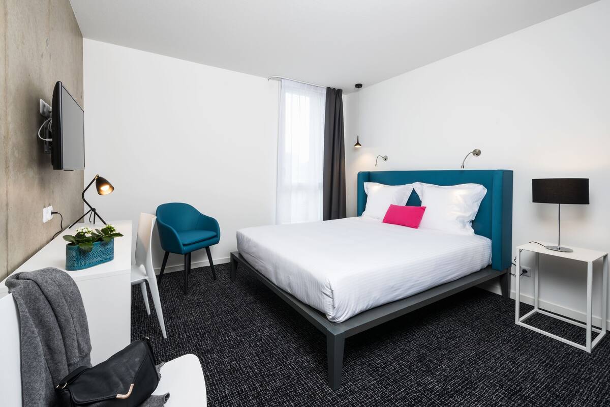 A modern hotel room with a bed, a desk, a chair, a TV, and a lamp.