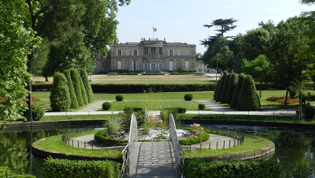 A large, historic building with a formal garden, a pond, and a bridge leading to the entrance.