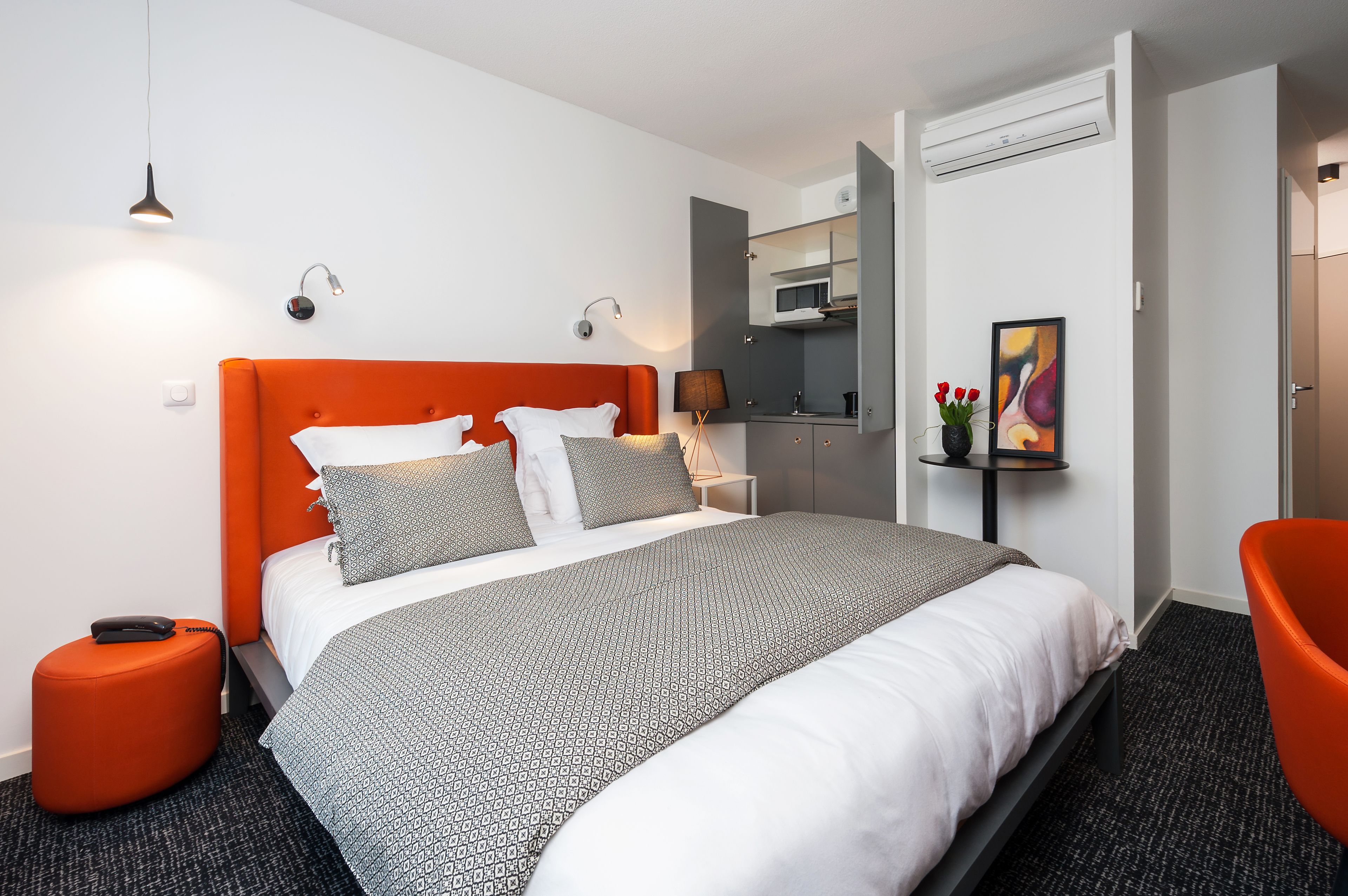 Habitació d'hotel moderna amb un llit gran, una paret color taronja, una petita zona de seients i una cuineta.