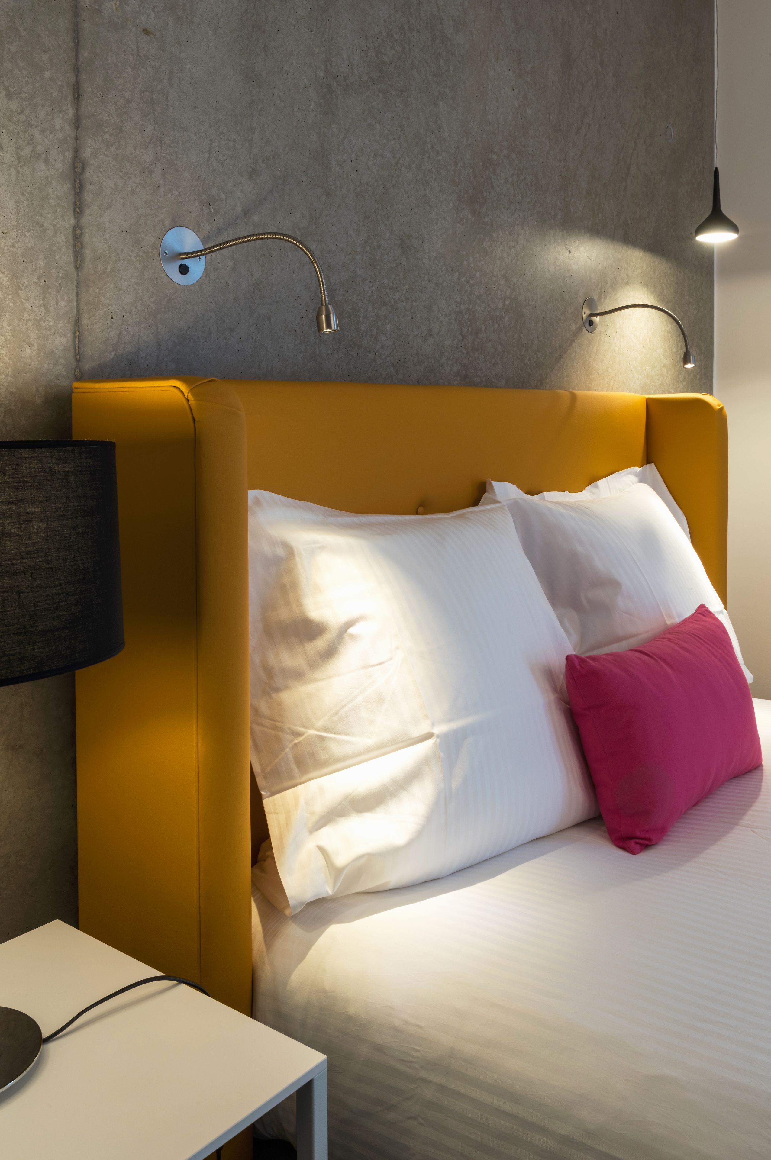 A modern bedroom with a yellow headboard, white and pink pillows, a bedside table with a lamp, and a wall-mounted light fixture.