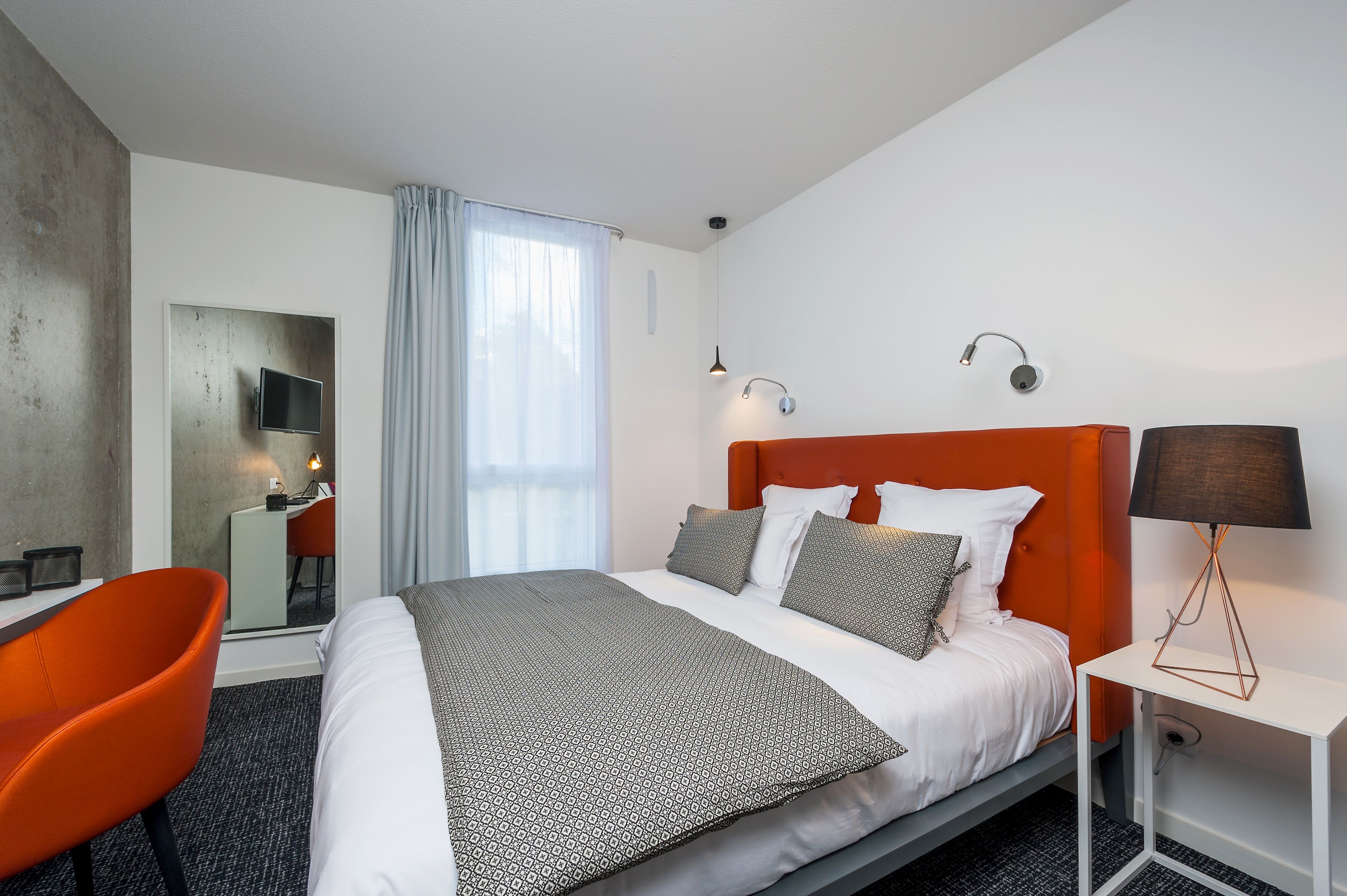 Une chambre d’hôtel avec un lit, un fauteuil orange, un bureau avec un ordinateur, un téléviseur fixé au mur et une lampe sur une table de chevet.