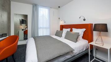 Une chambre d’hôtel avec un lit, un fauteuil orange, un bureau avec un ordinateur, un téléviseur fixé au mur et une lampe sur une table de chevet.
