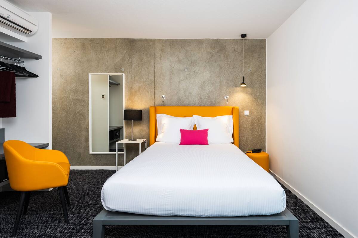 A hotel room with a bed, a chair, a bedside table, a lamp, and a mirror.