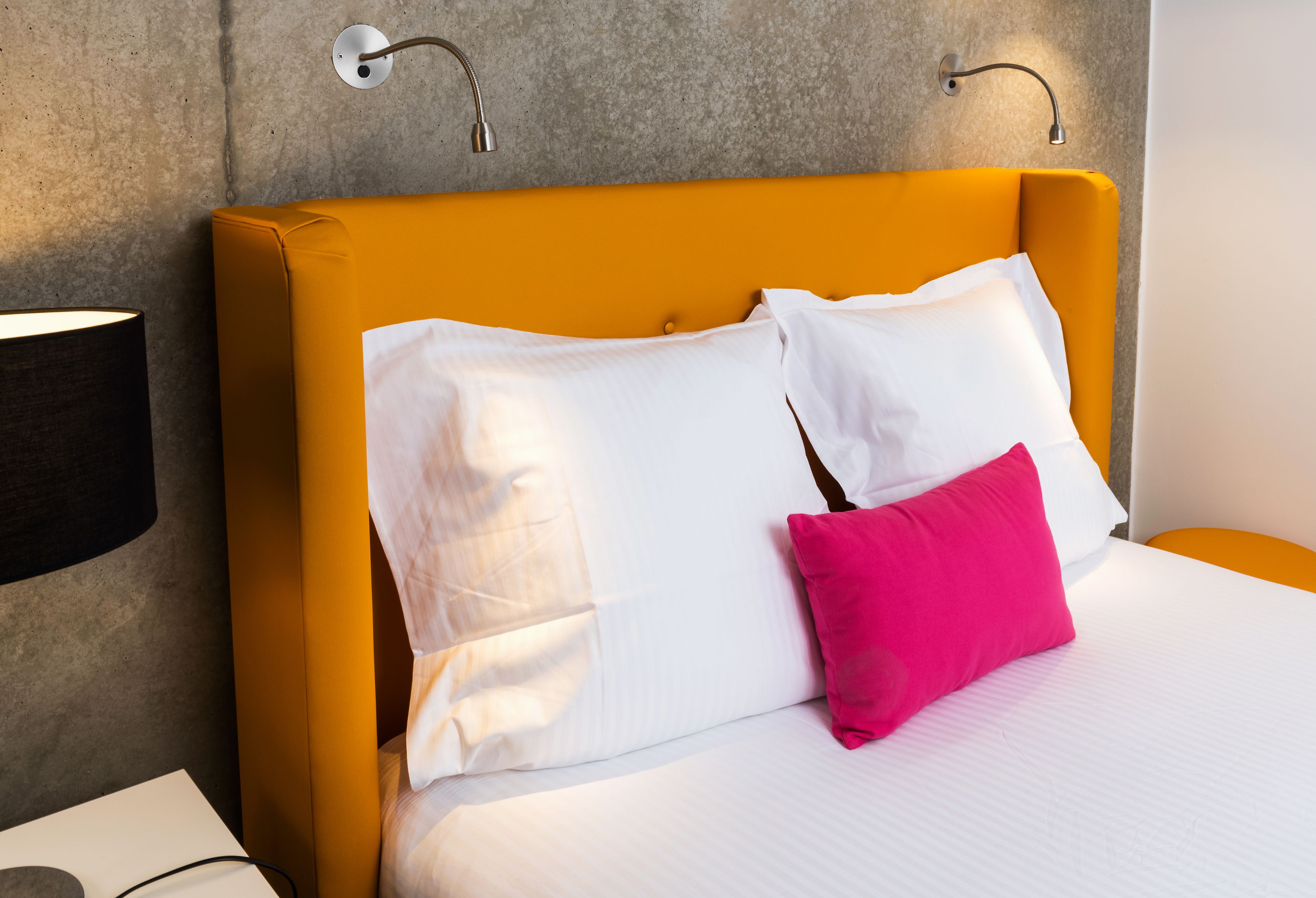Ein Hotelzimmer mit Bett, weißen und rosafarbenen Kissen, einer schwarzen Lampe und einer Wandleuchte.
