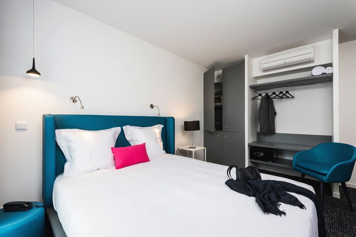 A hotel room with a bed, bedside table, lamp, wardrobe, and a chair.