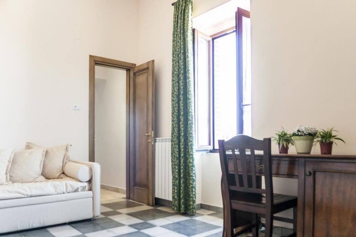 A room with a checkered floor, a white sofa, a wooden door, a window with green curtains, and a table with potted plants.