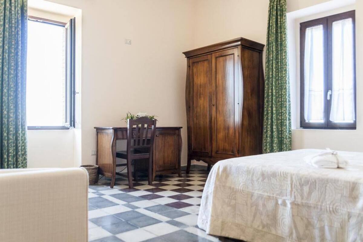 A room with a checkered floor, a bed with a white bedspread, a wooden desk with a chair, and a wardrobe.