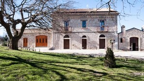 Un bâtiment en pierre doté de nombreuses portes et fenêtres en arc de cercle, entouré d’une zone herbeuse avec un grand arbre.