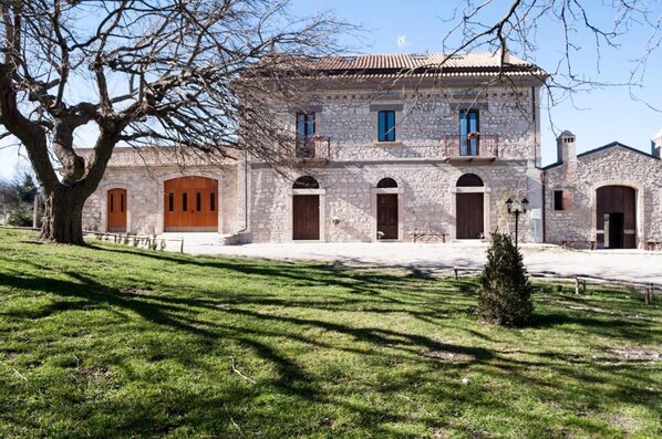 A stone building with multiple arched doorways and windows, surrounded by a grassy area with a large tree. - Masseria Salecchia (Bovino)