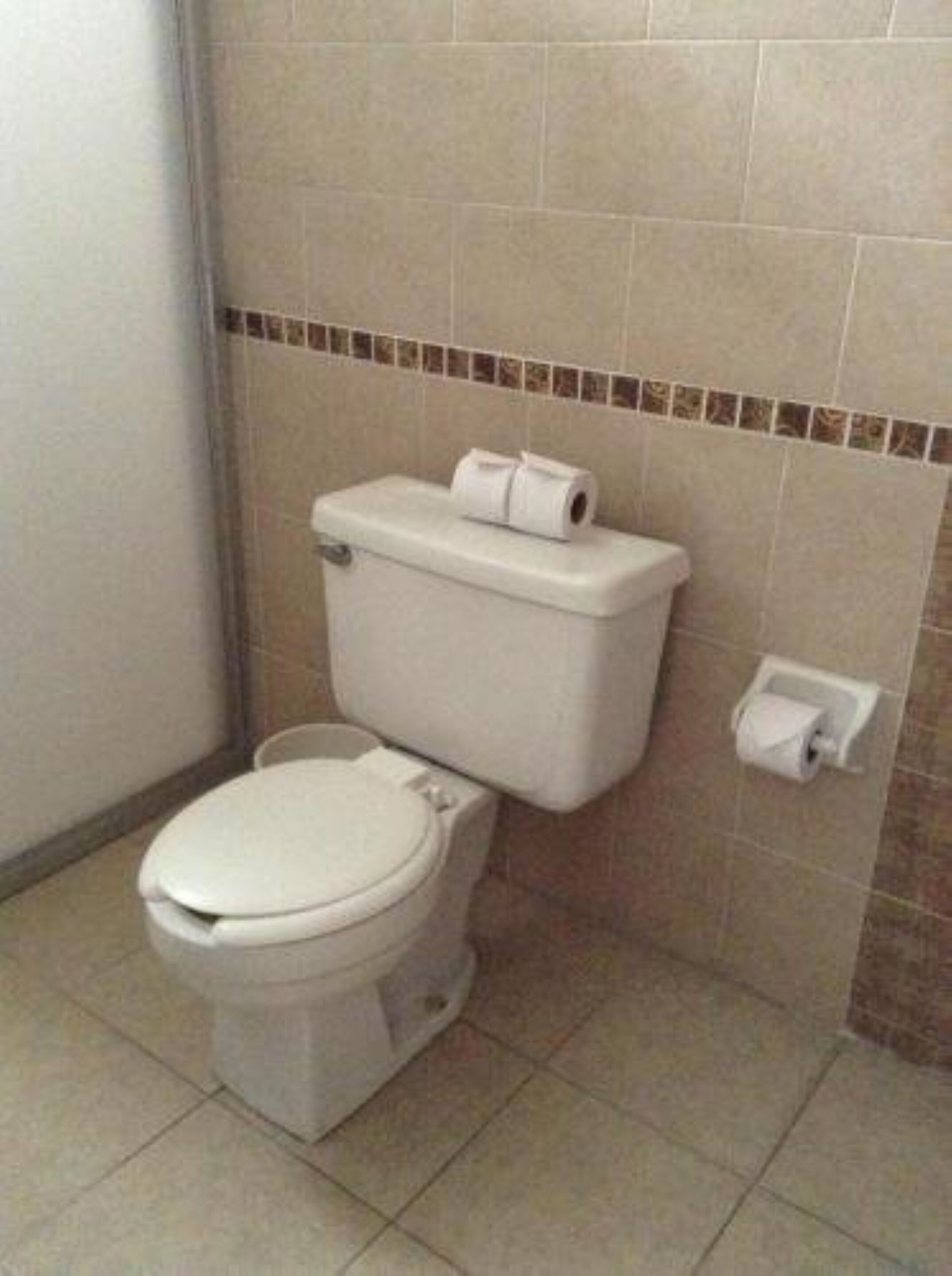 Un inodoro blanco estándar con un rollo de papel higiénico sobre el tanque y un dispensador de toallas de papel en la pared.