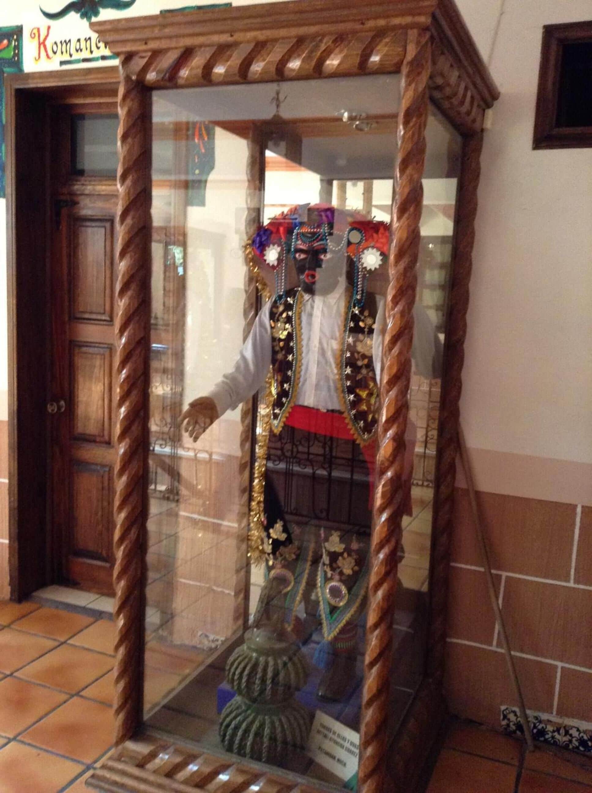 Un maniquí en una vitrina de vidrio con tocado ornamental, camisa blanca y falda roja.